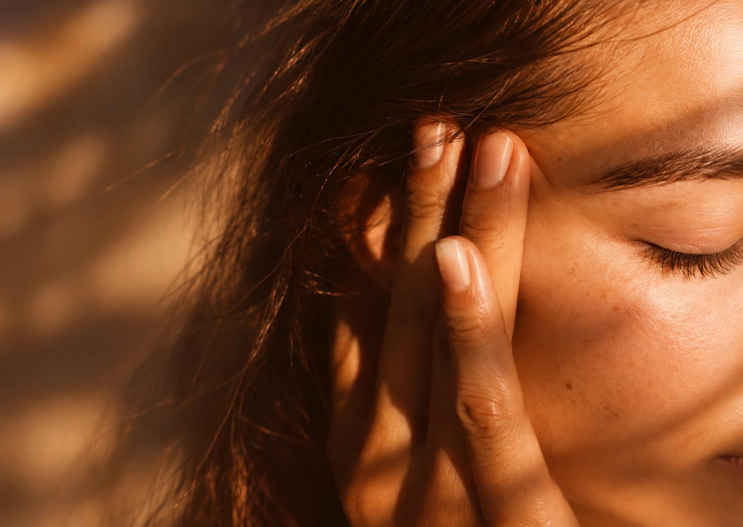 Femme les yeux fermés, doigts posés sur la tempe, dans une lumière chaude, évoquant les tensions que soulage un massage bien-être à Biarritz